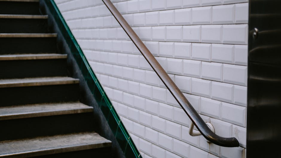 A downward view of a multi-story staircase with tiled steps and dark metal railings, featuring a central square light fixture on the wall and a hanging potted plant with green foliage and white flower