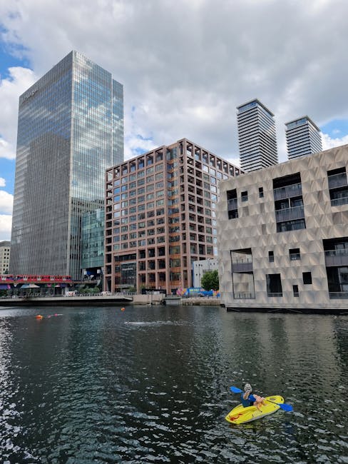 The image shows a person in a blue jacket and cap seated on a yellow kayak with a blue paddle, floating on the river near modern high-rise buildings in the Canary Wharf area. The buildings feature gla