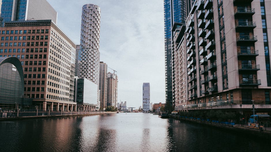 The image shows a person in a blue jacket and cap seated on a yellow kayak with a blue paddle, floating on the river near modern high-rise buildings in the Canary Wharf area. The buildings feature gla