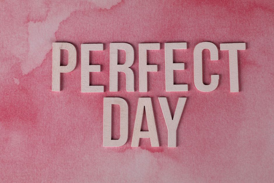 Close-up image of the words 'PERFECT DAY' spelled out in light-colored wooden letters placed on a pink watercolor painted surface. The letters are arranged in two lines, with 'PERFECT' on top and 'DAY